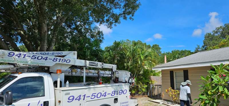 Bucket truck with 941-504-8190 on the boom parked at a home, a crew member in a sun hat walking toward the job.