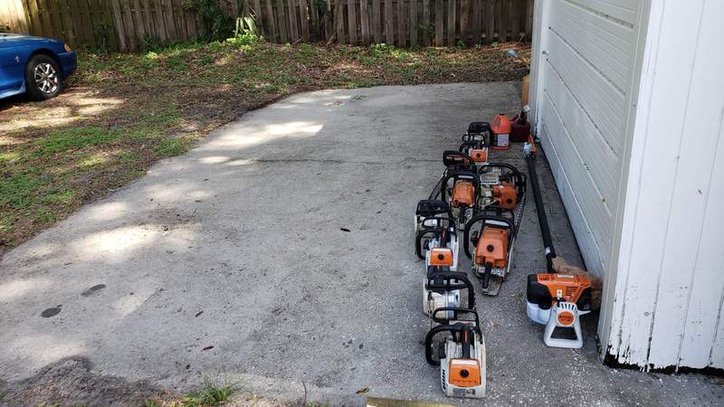 A row of orange-and-white chainsaws and a pole saw lined up on a concrete slab beside a shed.