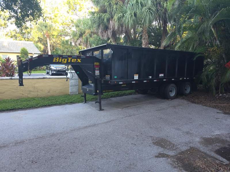 Black gooseneck dump trailer parked on a paved driveway in front of palms.