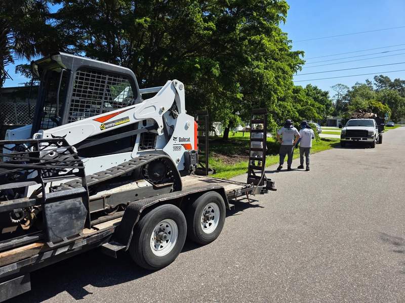 Bobcat track loader on a trailer at the curb, with two crew members talking by a dump truck down the street.
