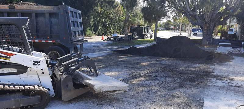 Track loader lifting a broken slab of old concrete, with a dump truck and a pile of fill dirt behind it.